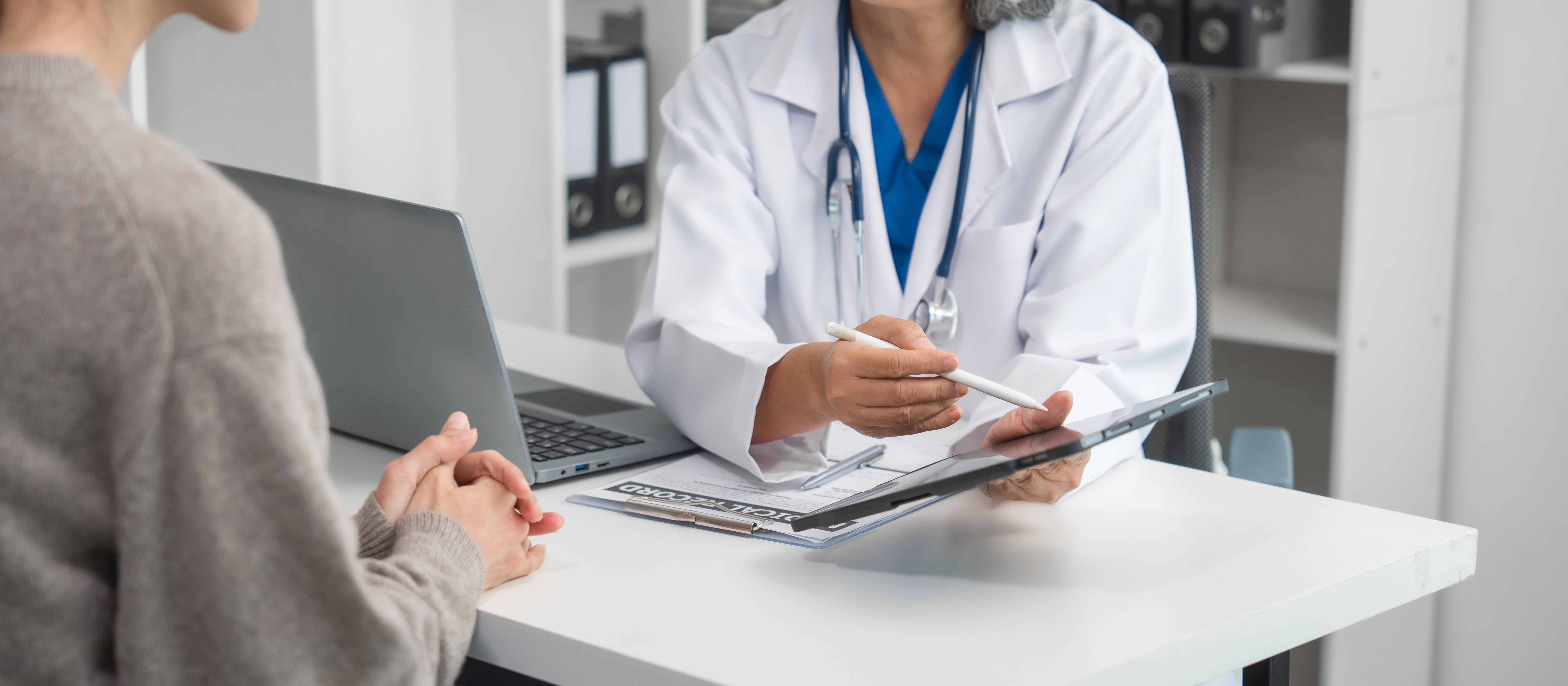 Ärztin spricht mit einer Patientin am Schreibtisch und hält dabei ein Tablet.