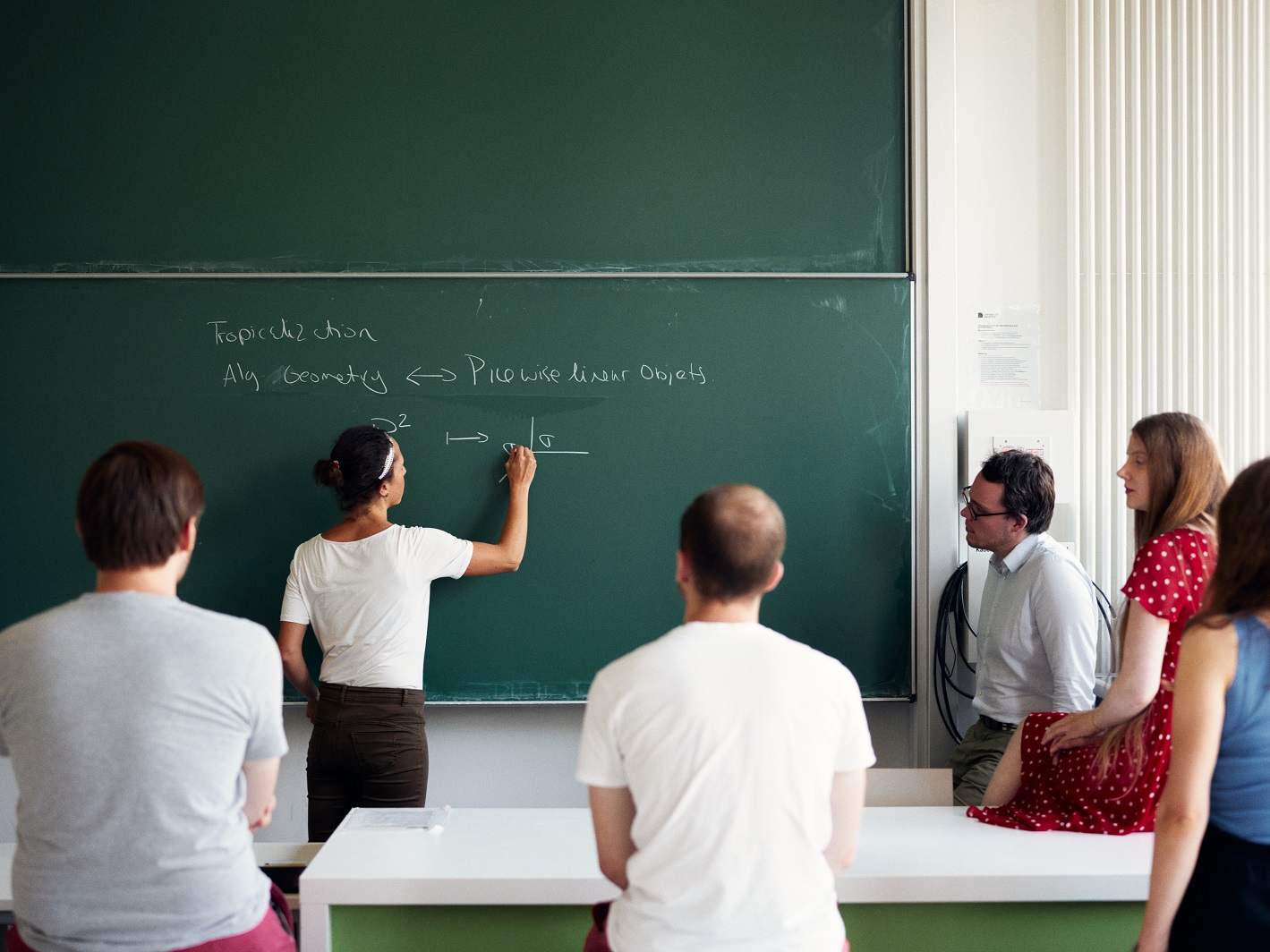 Fünf Personen sitzen vor einer Tafel und sehen zu, wie eine Person mathematische Formeln anschreibt