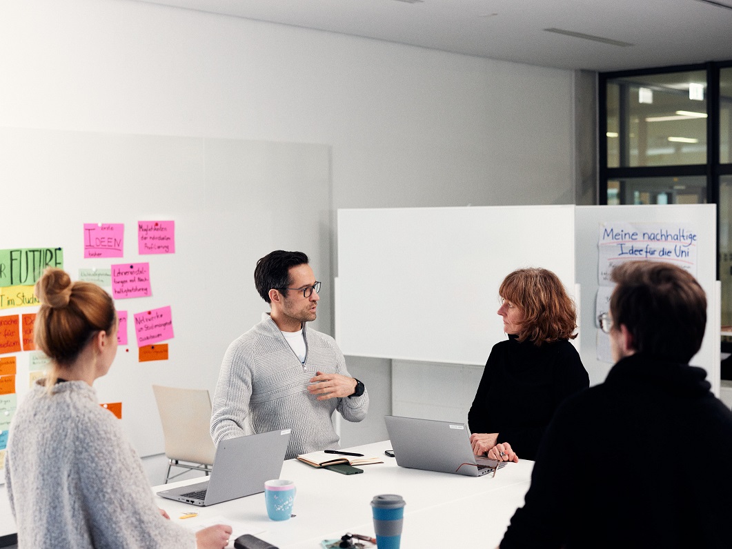 Eine Gruppe von vier Personen sitzen um einen Tisch. Hinter ihnen befinden sich Pinnwände, an denen bunte Karten hängen. 