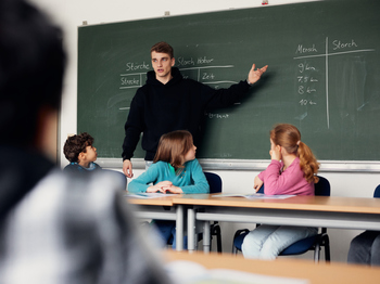 Lehrkraft steht an der Tafel und erklärt Grundschulkindern etwas, indem er mit der Hand auf die Tafel zeigt.