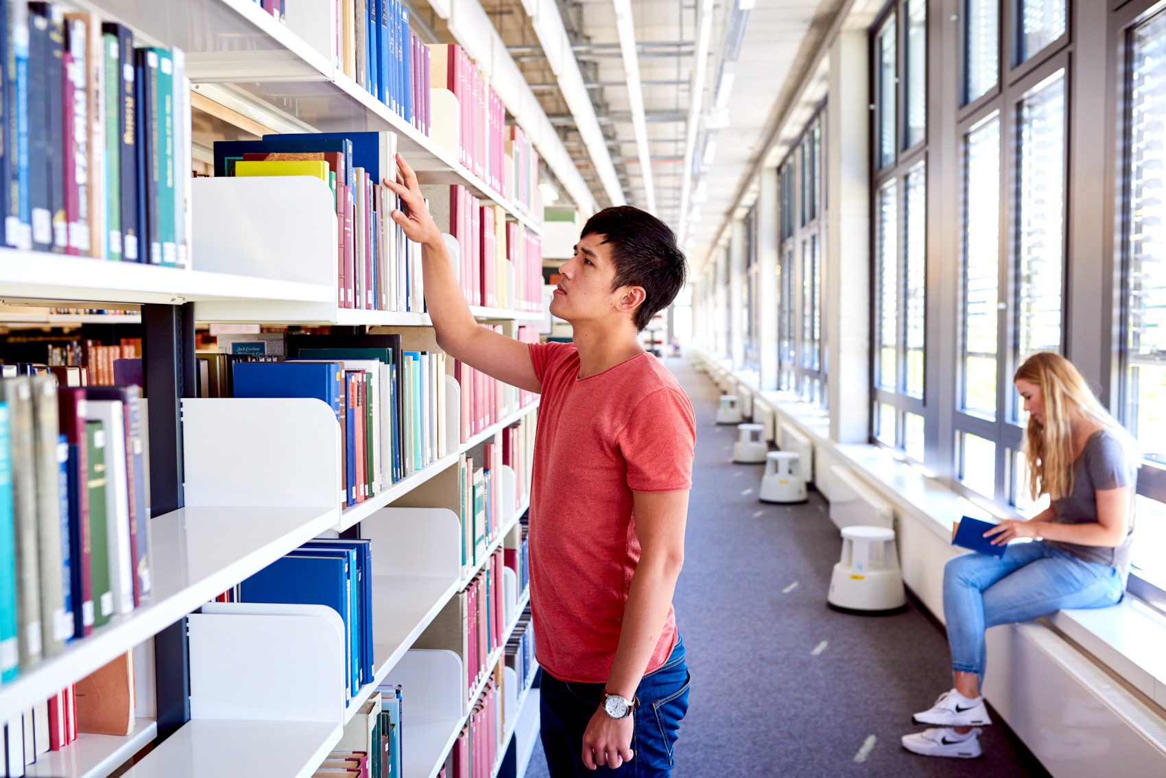 Studierende in der Bibliothek, Student an einem Regal, eine Studentin sitzt und blättert in einem Buch