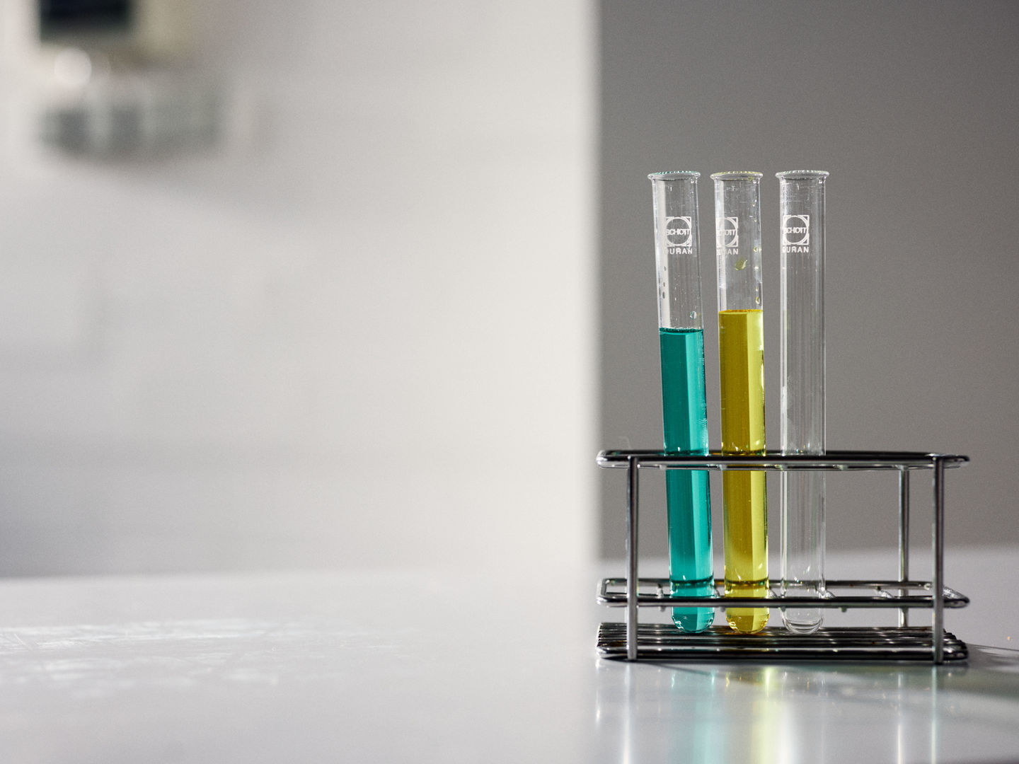 Drei Reagenzgläser in einem Reagenzglasständer. Von links: Türkise Flüssigkeit, gelbe Flüssigkeit, leer.