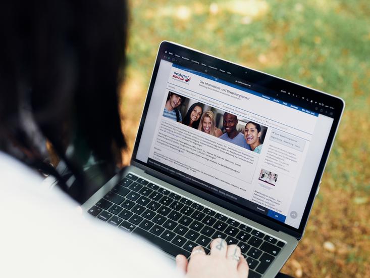 Overshoulder Shot über die Schulter einer schwarzhaarigen Person, die auf einen Laptop blickt. Auf dem Bildschirm ist die Website der Studienplatzvergabe zu sehen.