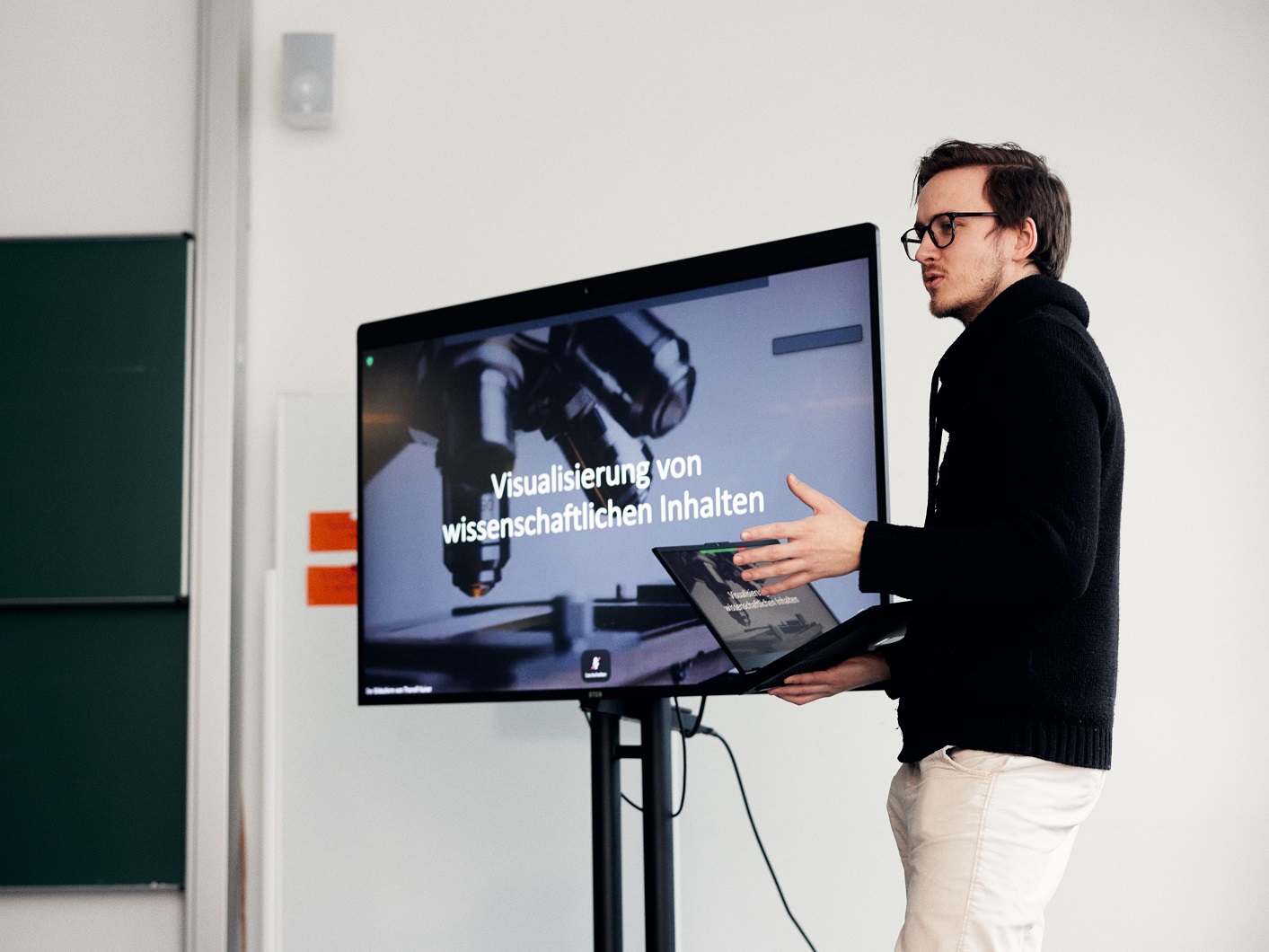 Ein Mann hlt einen Laptop und stellt eine PowerPoint-Prsentation vor, die neben ihm auf einem groen Bildschirm angezeigt wird